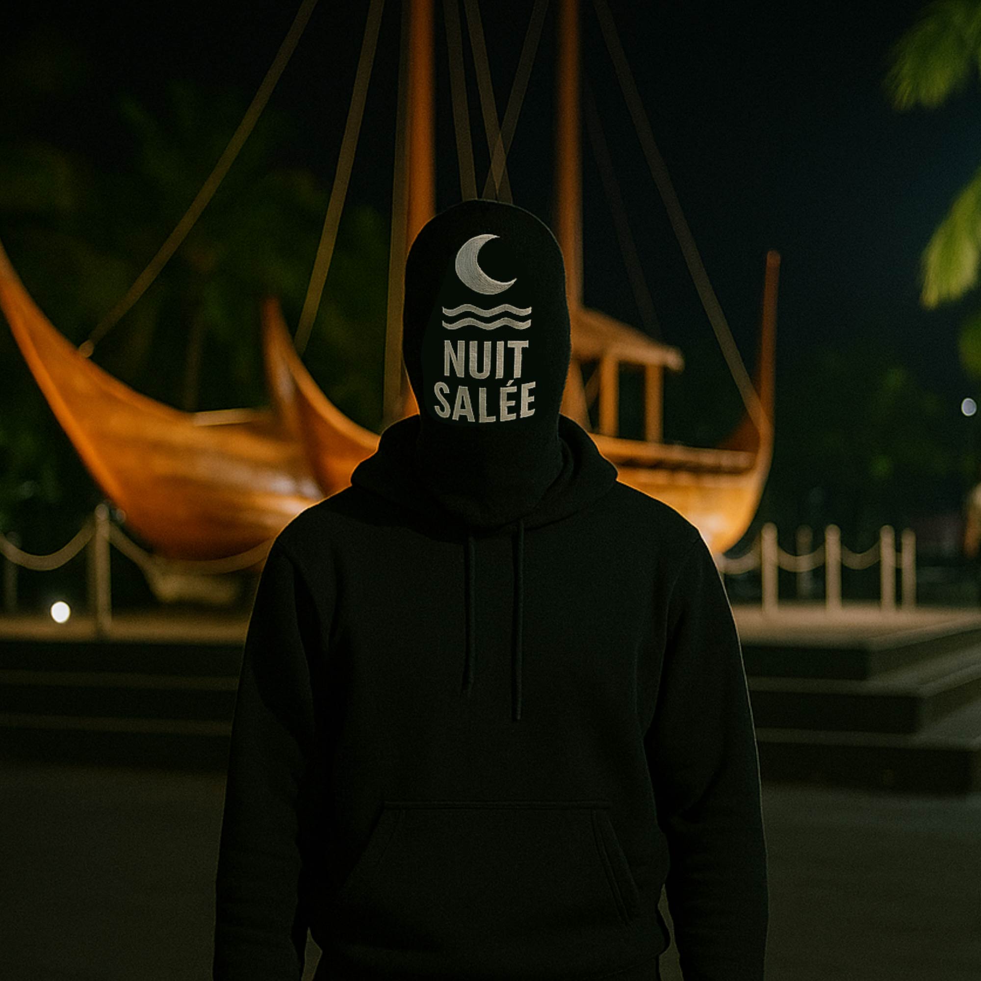 Nuit Salée in a black hoodie in front of a traditional sailing canoe at night.